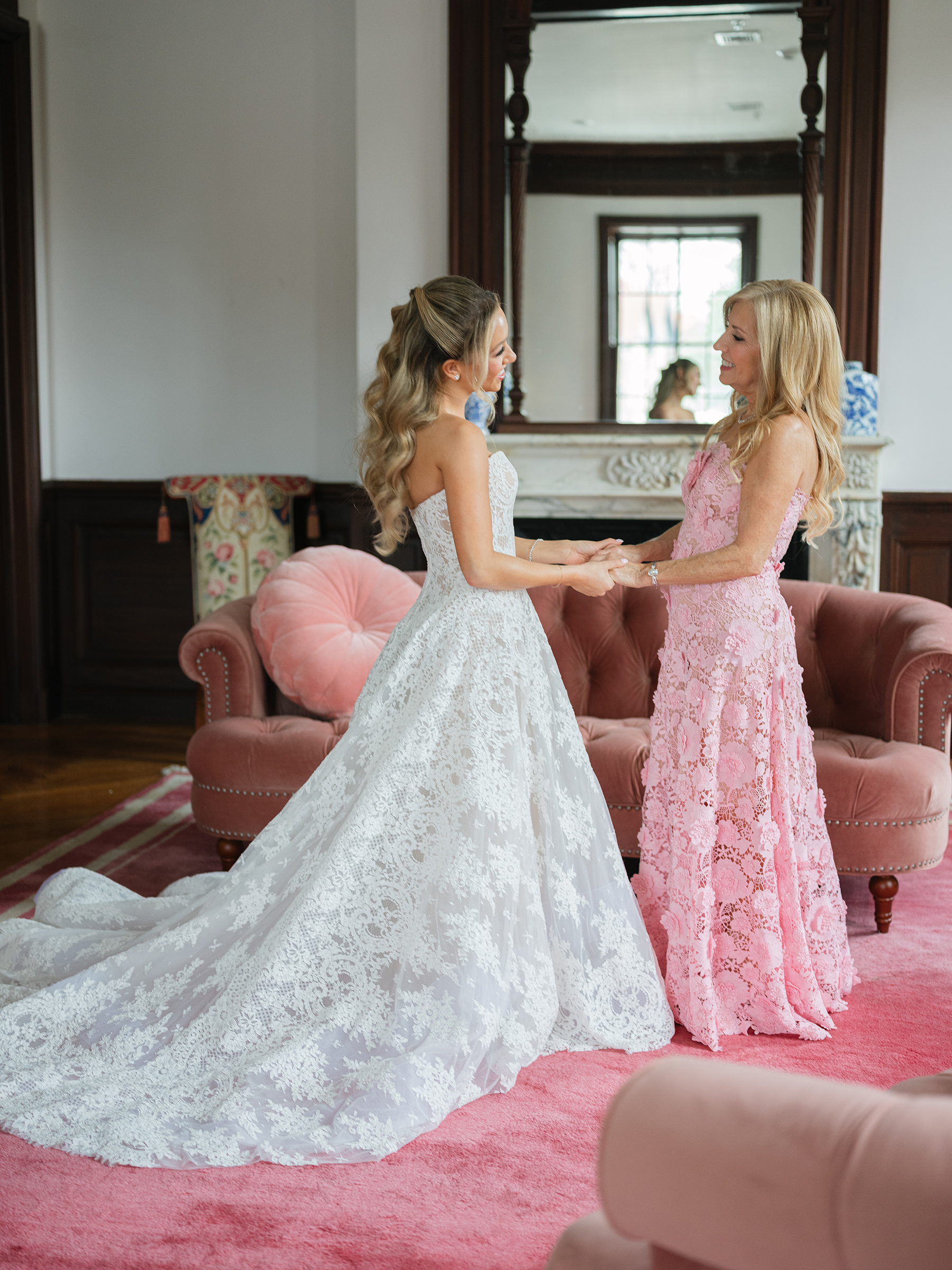 bride wearing a corset-top dress with long train, holding hands with her mom in a baby pink lace dress