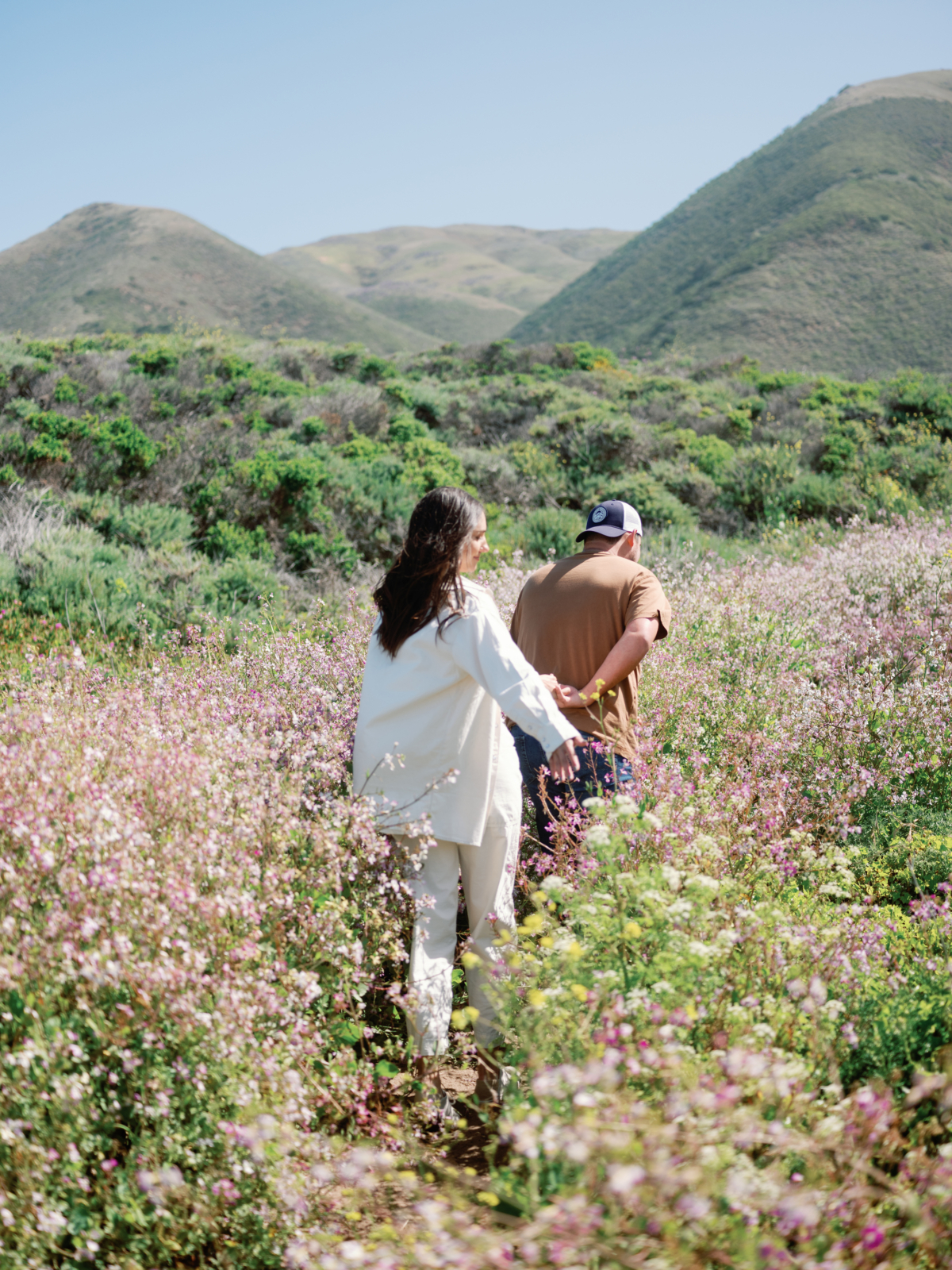 Couple walking through pink wildflowers toward rolling green hills near Carmel, California.
