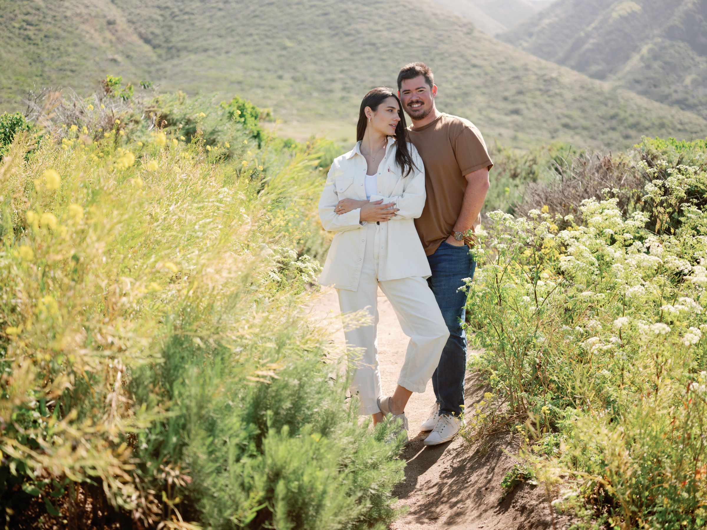 Couple standing close on a hillside trail lined with wildflowers near Carmel.