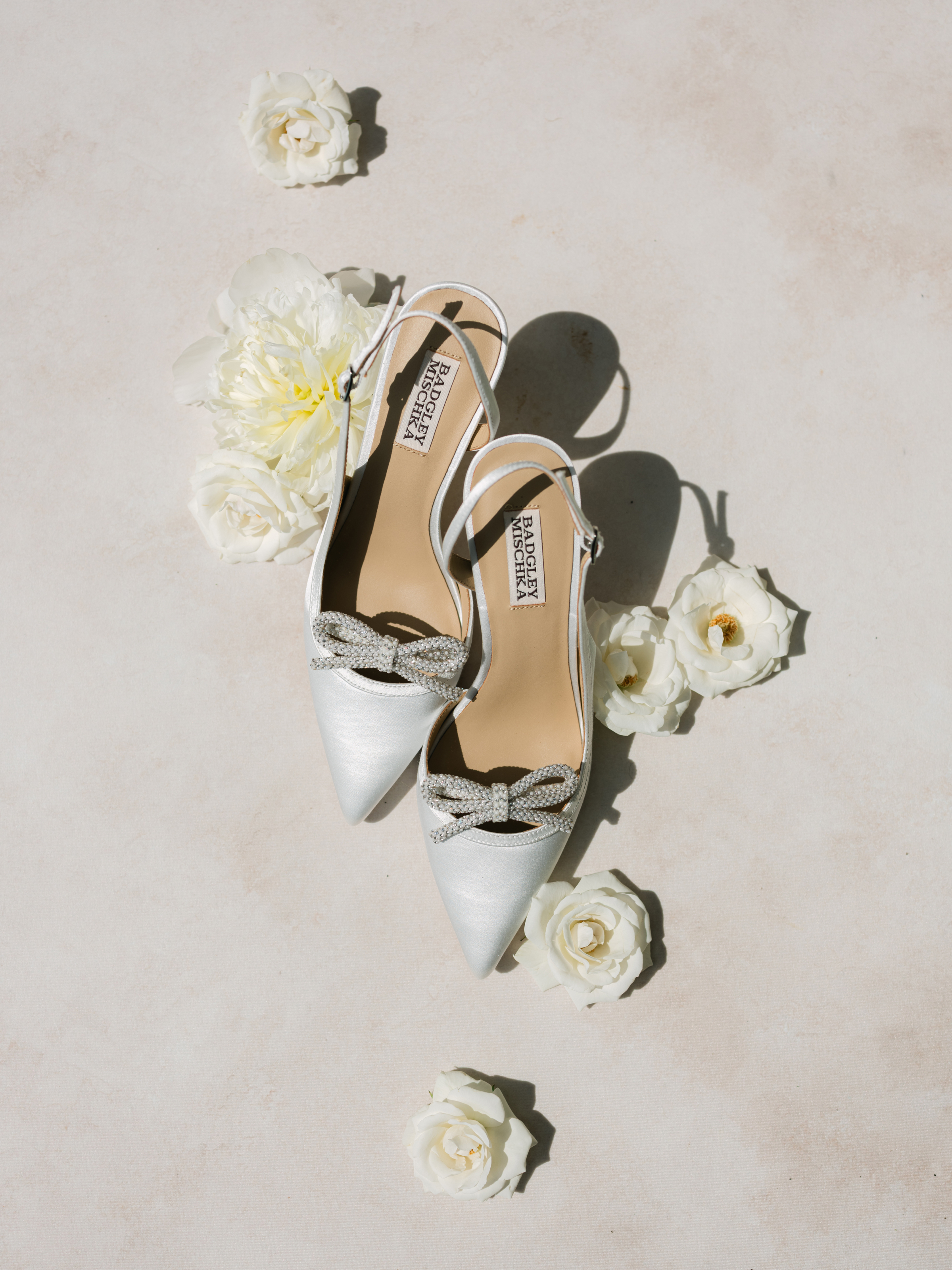 Ivory satin Badgley Mischka slingback heels with crystal bows laid among white roses.