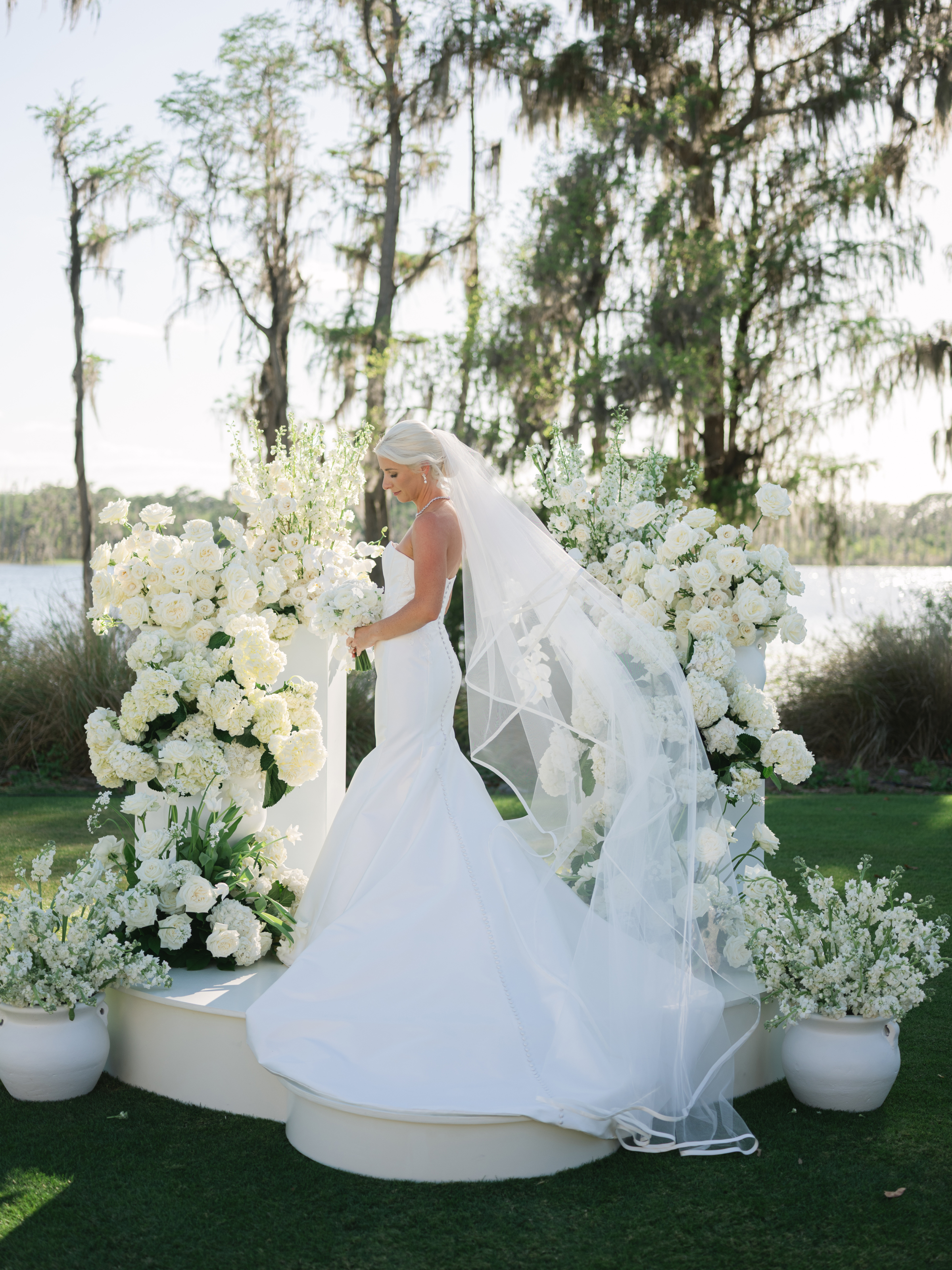 Hannah stands on the ceremony stage with her cathedral veil and gown train spread around her and the lake behind.