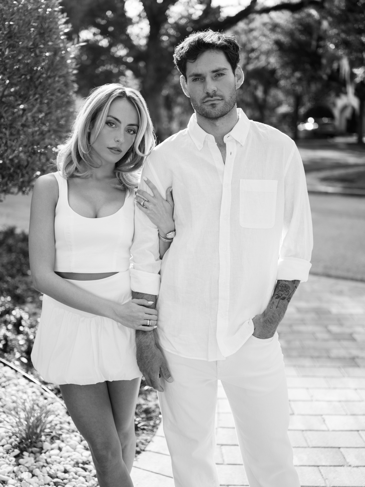 Black and white portrait of Lauren and Joao in matching all-white outfits on an oak-lined street in Winter Park.