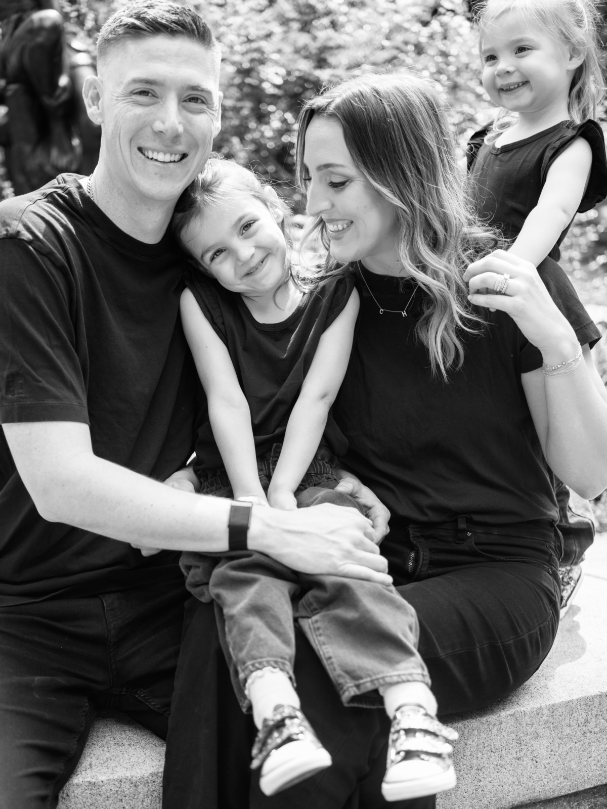 Black and white photo of the family cuddled together on a garden wall, the girls grinning at the camera.