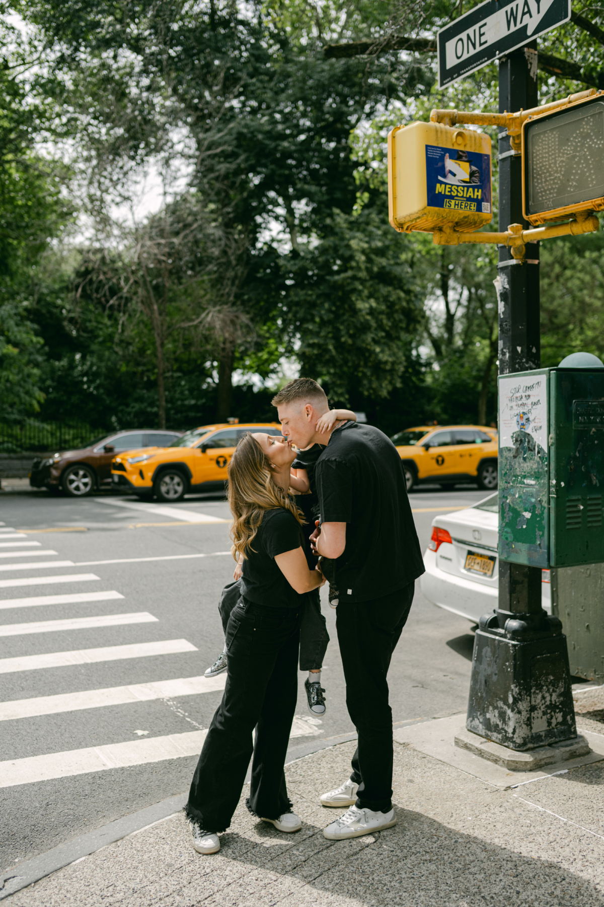 Nicole and Ryan kiss on a street corner under a One Way sign with yellow cabs behind them.