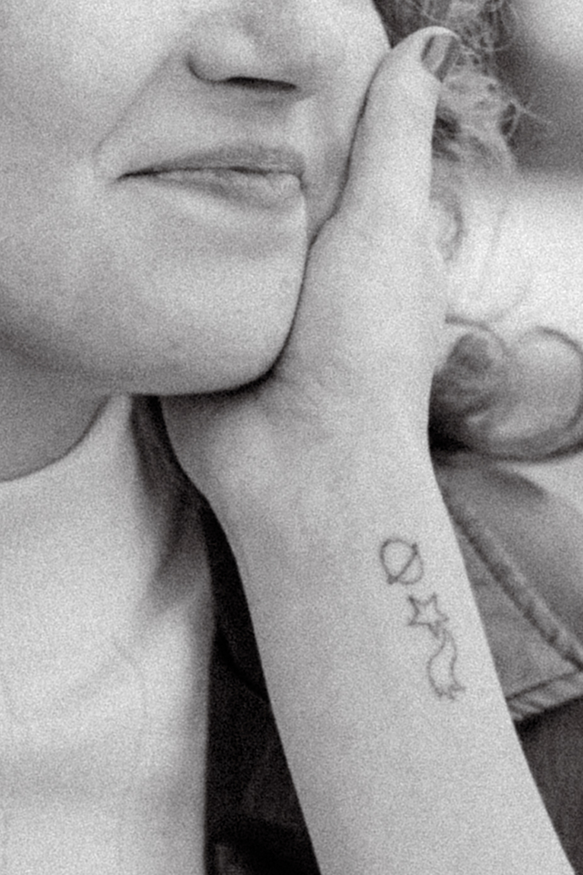 A black and white detail of a hand on a cheek, showing a small planet, star and moon wrist tattoo.